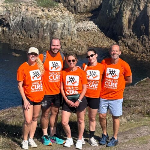 A group of people in orange Walk to Cure Parkinson’s t-shirts pose in front of the ocean, with a clear blue sky and waves in the background.